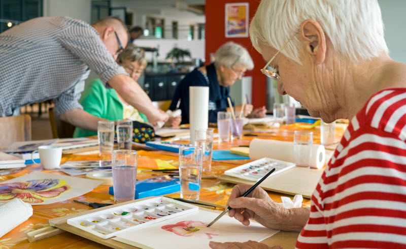 Woman in striped red and white shirt working on watercolor painting at table with other students