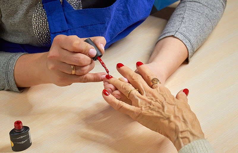 Manicurist painting nails to elderly woman with red polish on salon table.