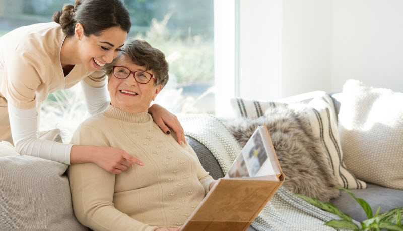 Grandmother sitting on a couch in a sunny room, looking at a photo album and sharing fond memories with a tender caregiver