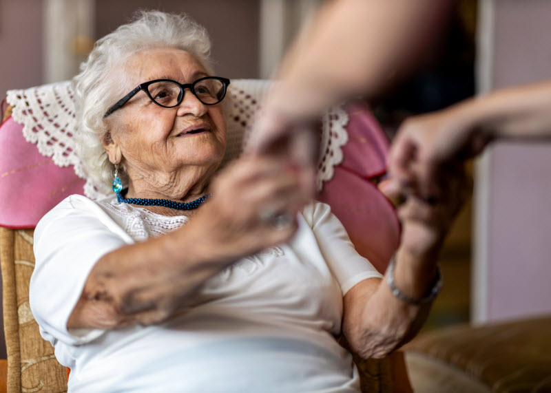Female home carer supporting old woman to stand up from the armchair at care home