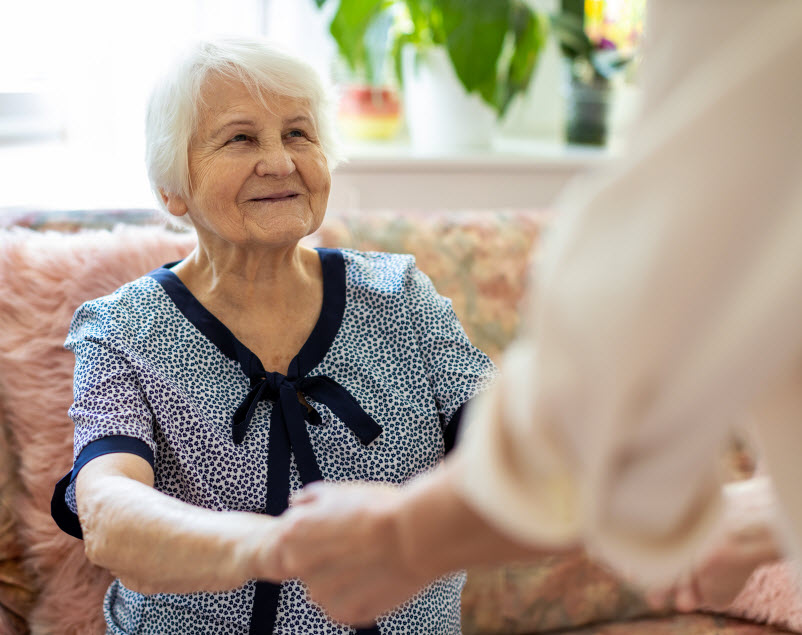 Female home carer supporting old woman to stand up from the sofa at care home